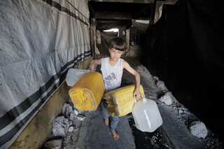 A barefoot boy in a white singlet carries four empty water containers through a rubble-filled corridor