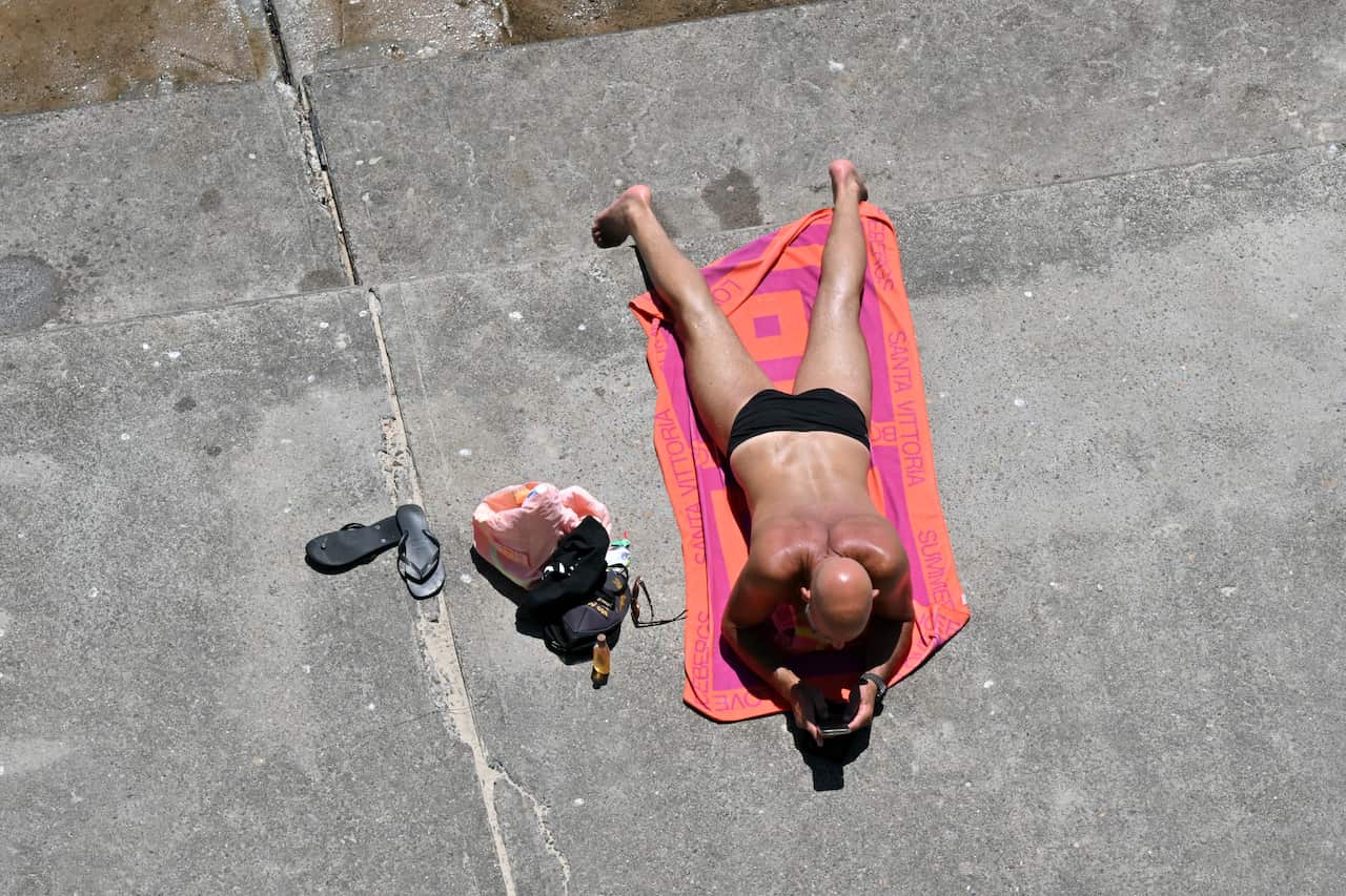 A man in his swimmers lies face down on a towel on a slab of concrete amid the summer heat.
