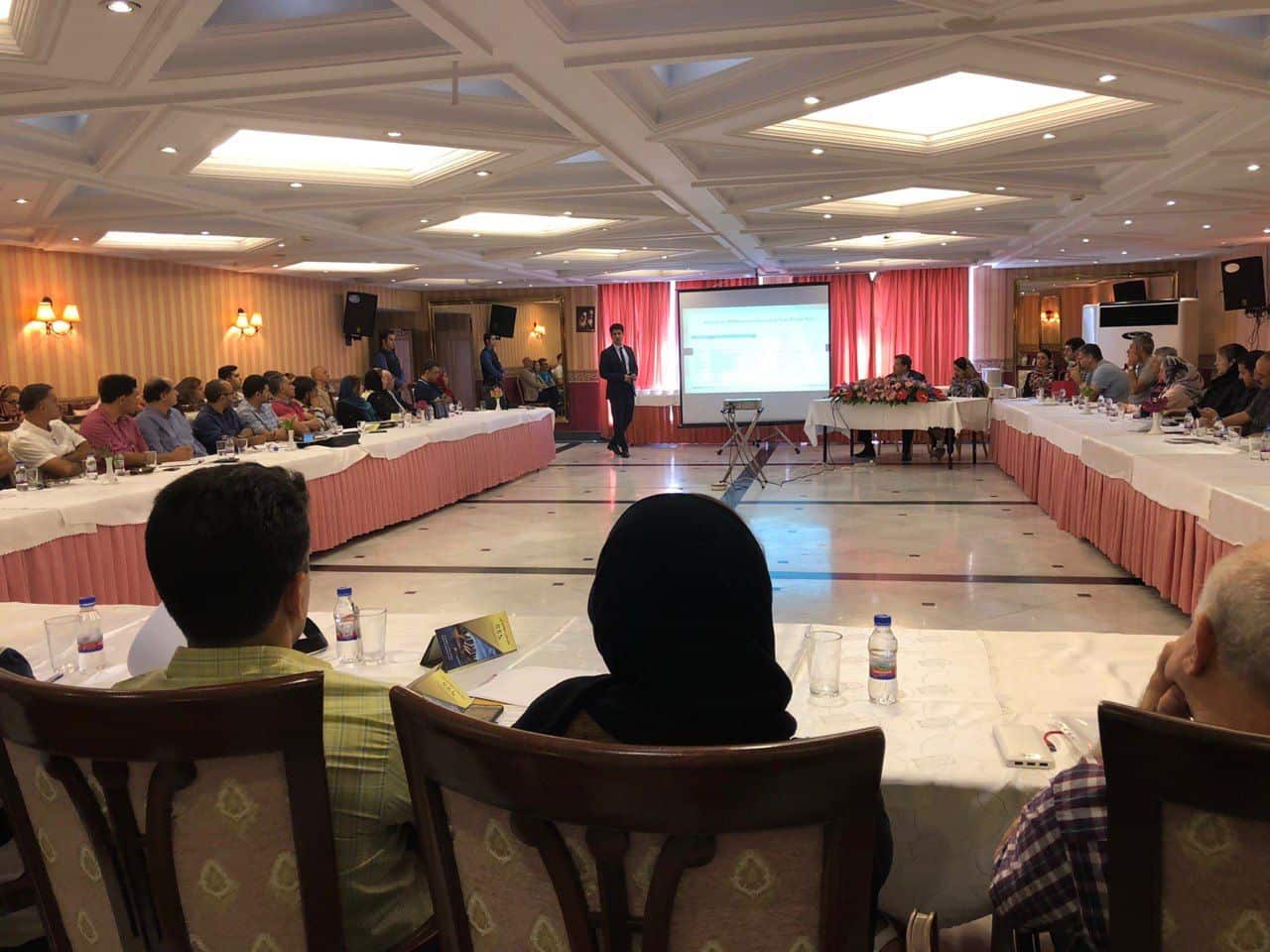 A seminar presenter in a suit stands on a stage between two long tables, addressing an audience. The attendees are seated and facing a large projection screen at the front of the room.