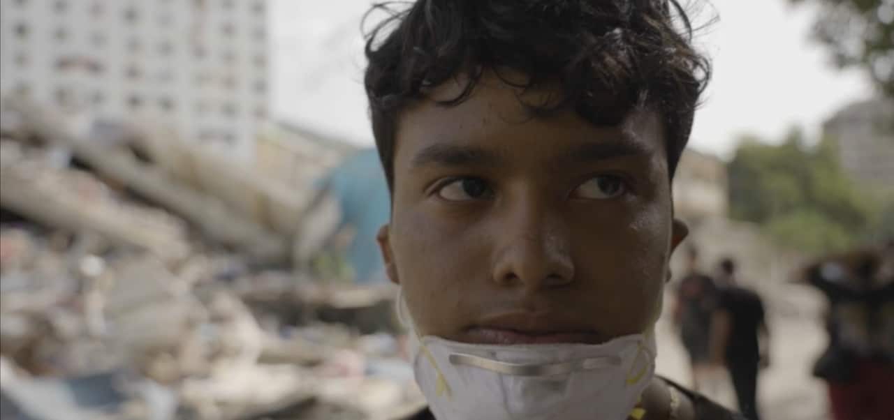 A close-up of a young boy with a face mask pulled down. There is rubble of collapsed buildings in the background.