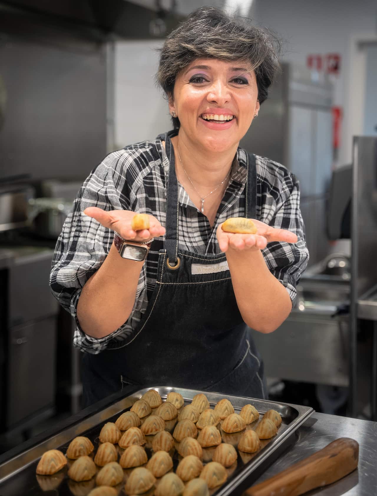 A woman in a checked shirt holds maamool in a commercial kitchen.