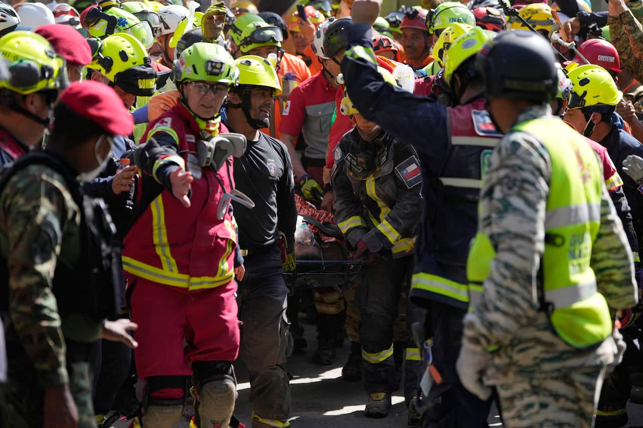 A crowd of rescuers wearing red, yellow and camouflage print surround a stretcher. Some have their arms raised in celebration.