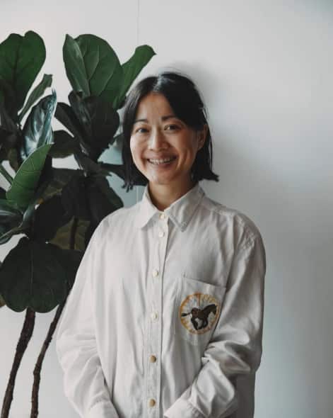 A woman in a white shirt smiling in front of a pot plant.
