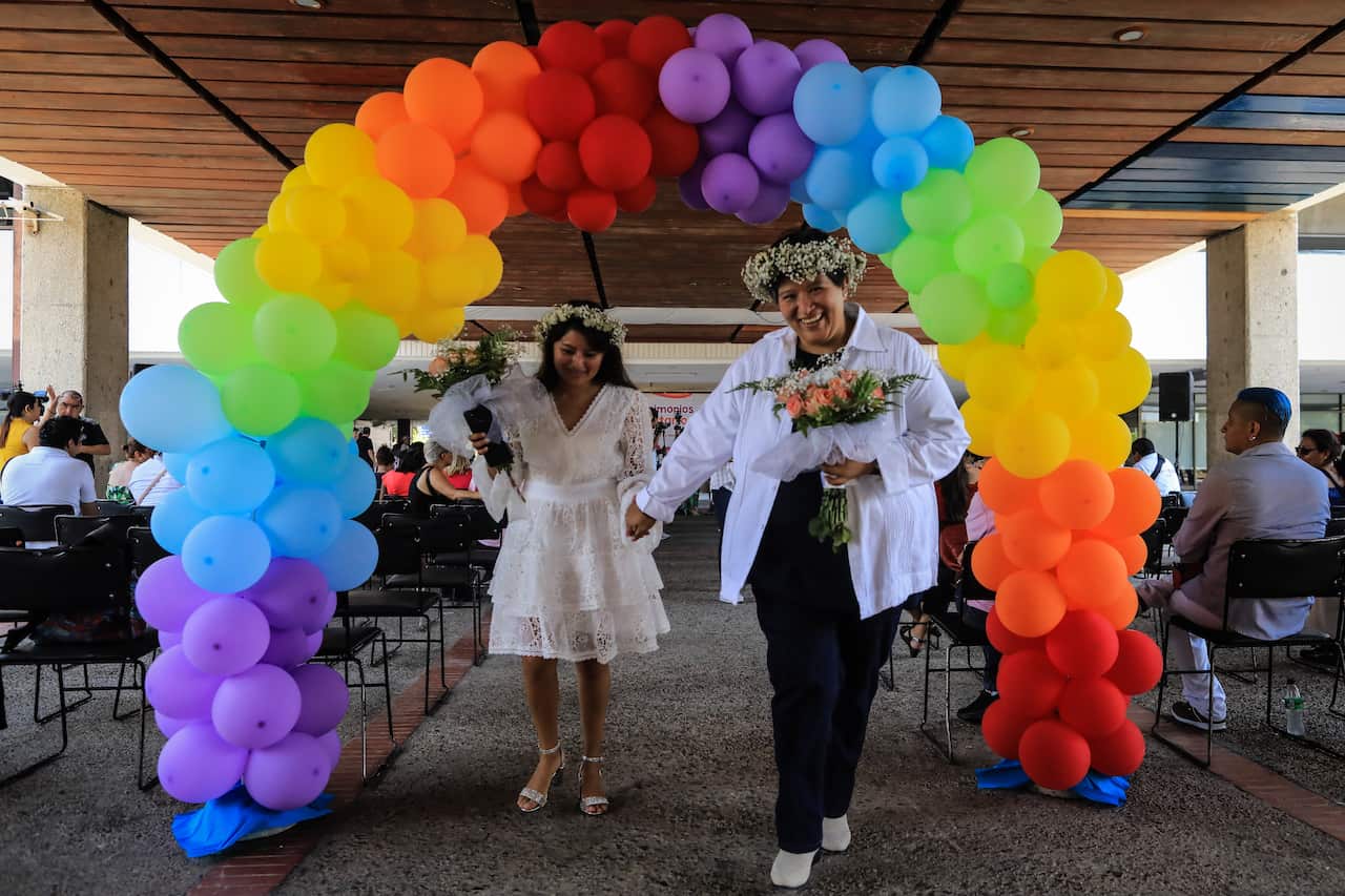 A newly wed couple walk under rainbow balloons.