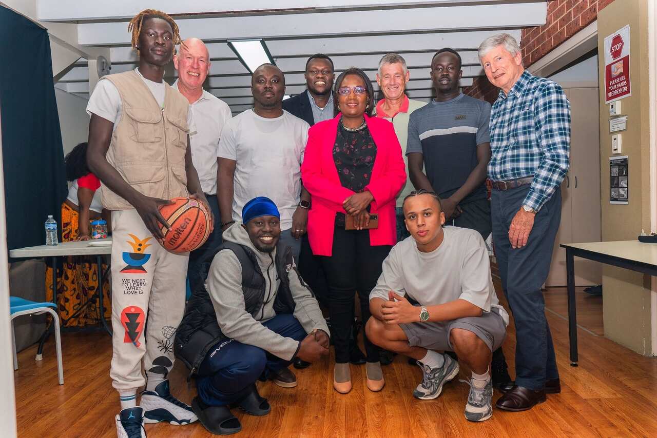 A woman in a fuscia blazer poses with several young African-Australian men and several older caucasian men.