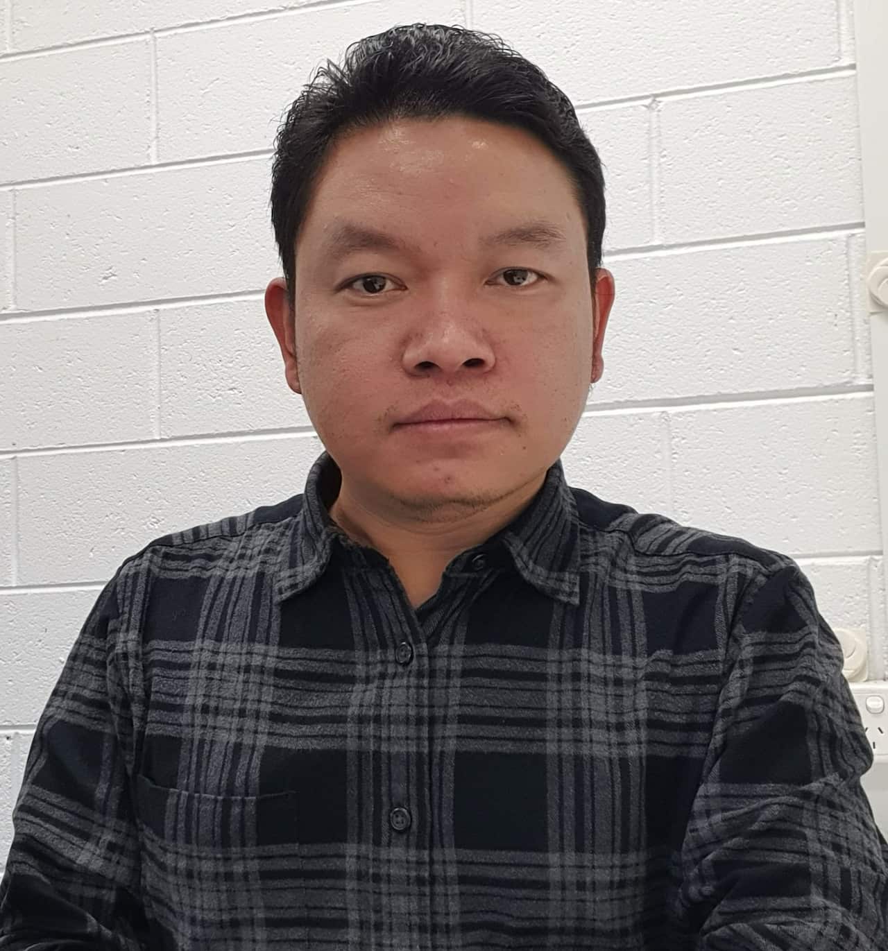 James looks at the camera with a serious facial expression, wearing a dark checkered button-up shirt.