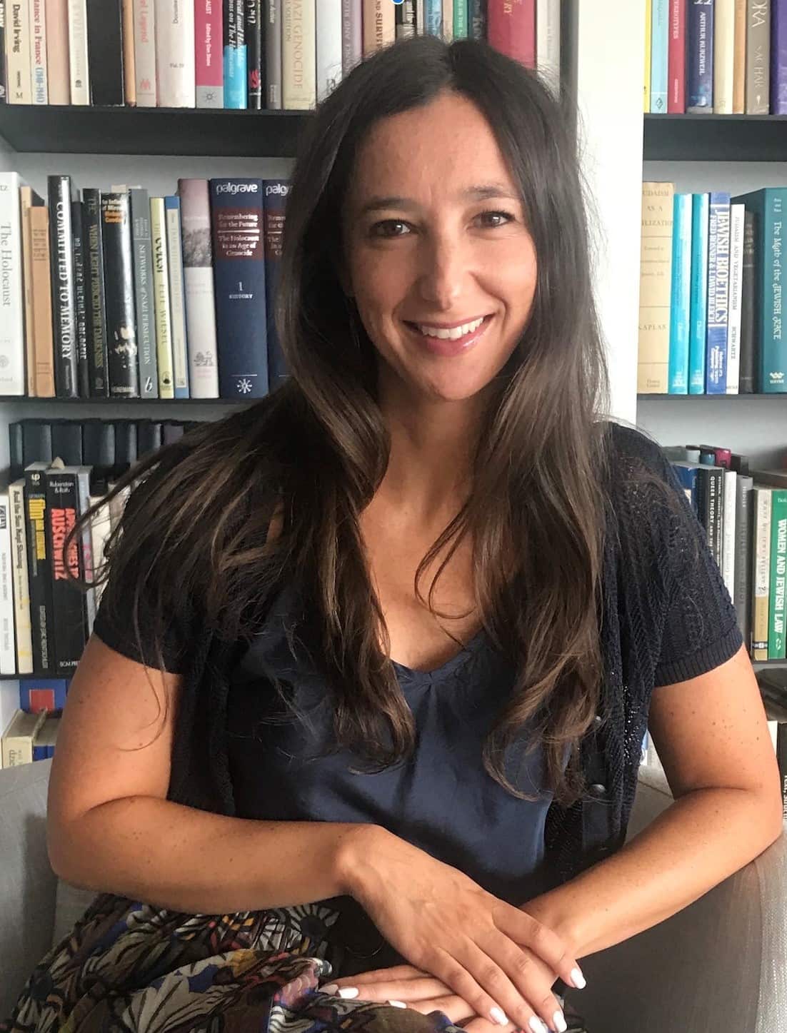 A woman with long brown hair sits in front of a bookshelf looking forward and smiling.