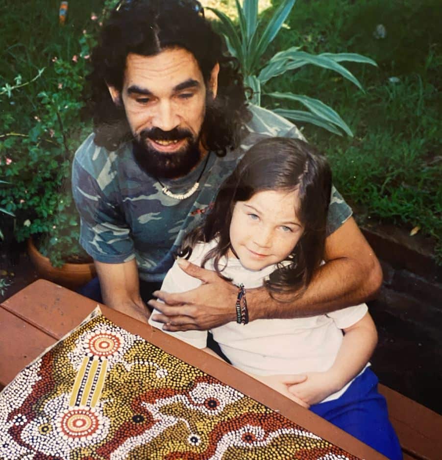 A young girl with her dad ,who he embraces on his lap. There is an Indigenous painting on the table in front of them.