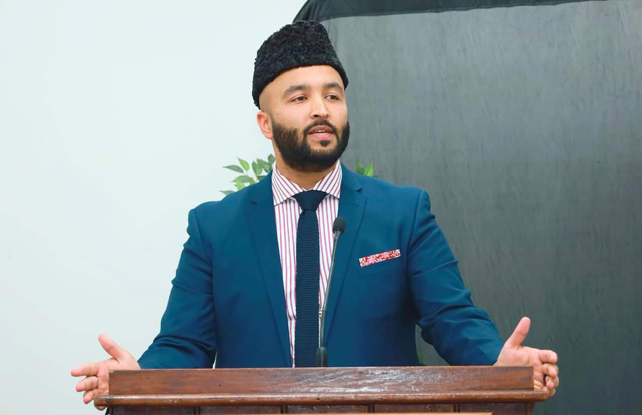 A man stands at a lectern wearing a blue suit, red and white striped shirt, and a blue tie