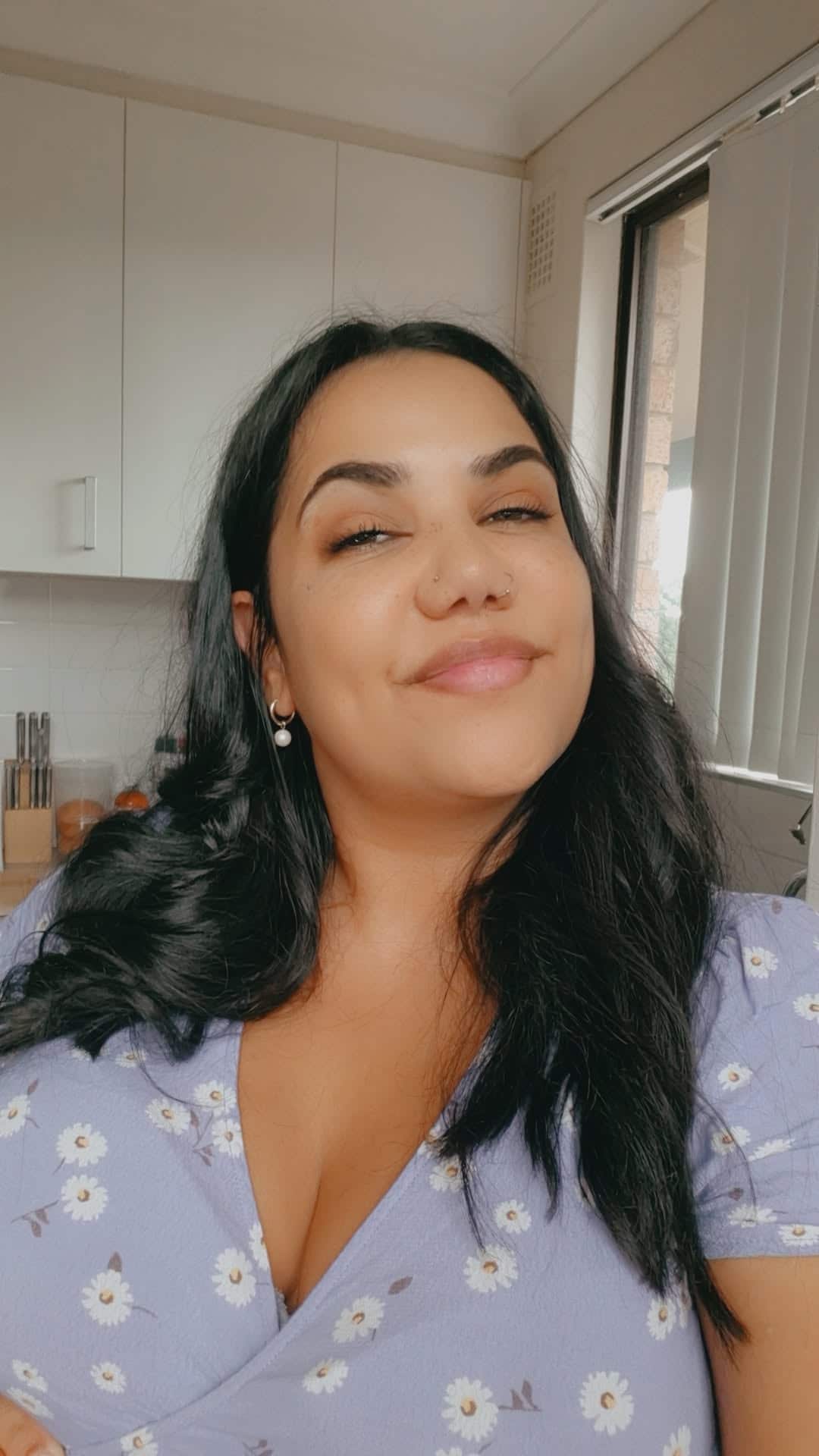 A selfie of a woman in a blue floral dress with long dark hair and a smile on her face.
