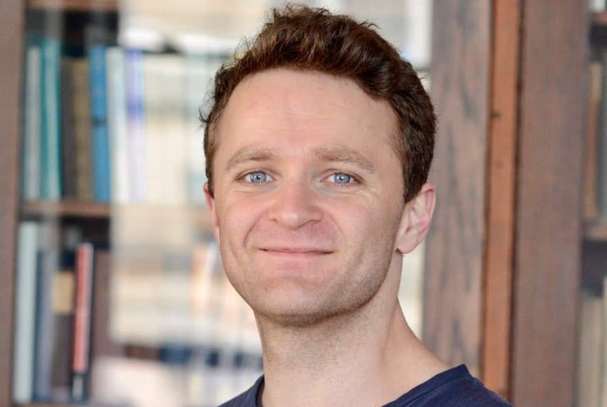 A close-up photograph of 33-year-old Alistair Kitchen. He smiles with pursed lips and stands in front of a bookcase, blurred in the background.