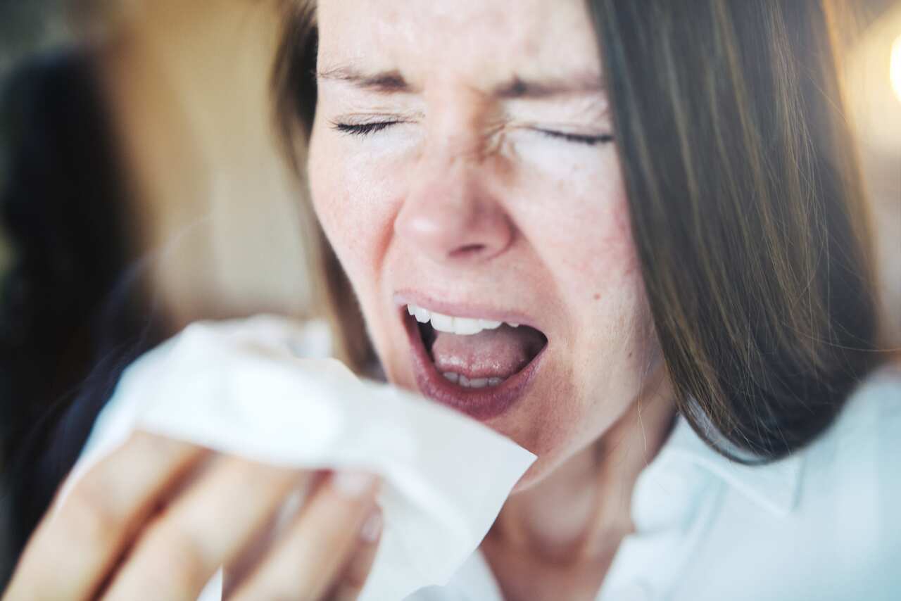 Woman sneezing loud with open mouth behind a window.
