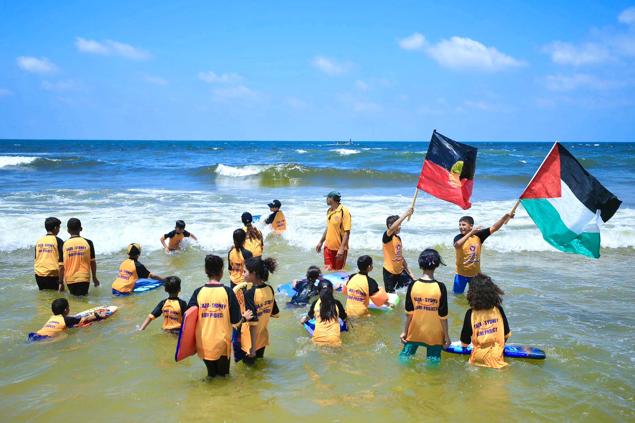 A group of children and one adult stand in the ocean surf on a bright, sunny day. The children are wearing matching orange shirts and are waving two flags: a Palestinian flag and Aboriginal Australian flag.