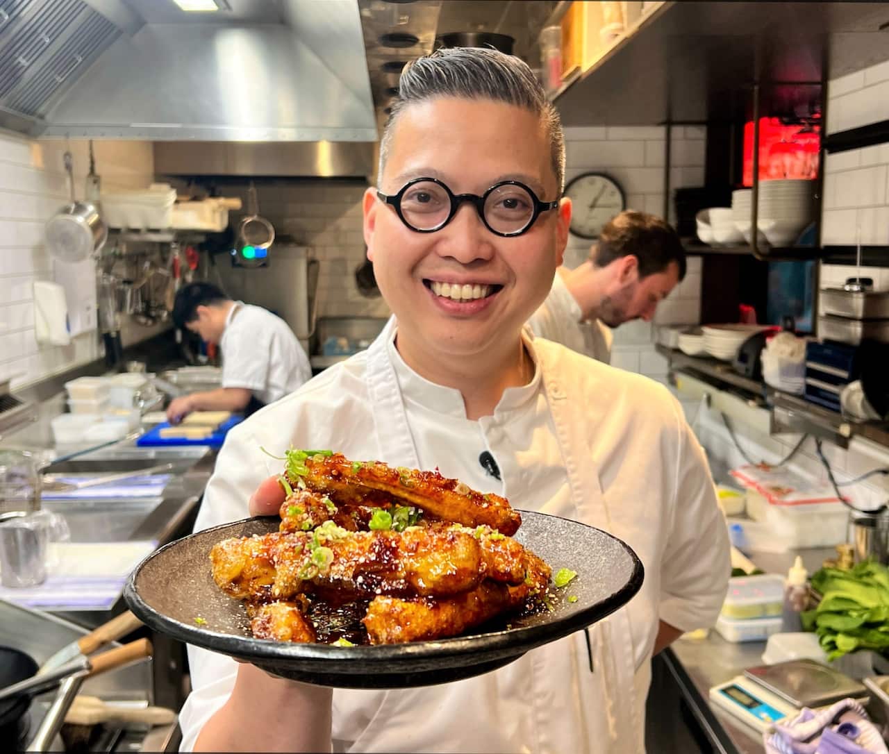 A man wearing round glasses and a white chef uniform holds a plate of fried food.