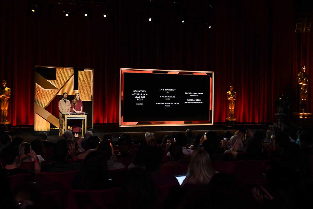 Two people in a theatre on stage behind a lectern. A large screen next to them displays peoples' names. There is a seated audience in the theatre. 