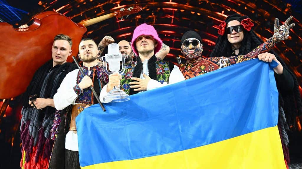 Six members of a band pose behind the Ukrainian blue and yellow flag. One member is holding a glass trophy which is shaped as a microphone