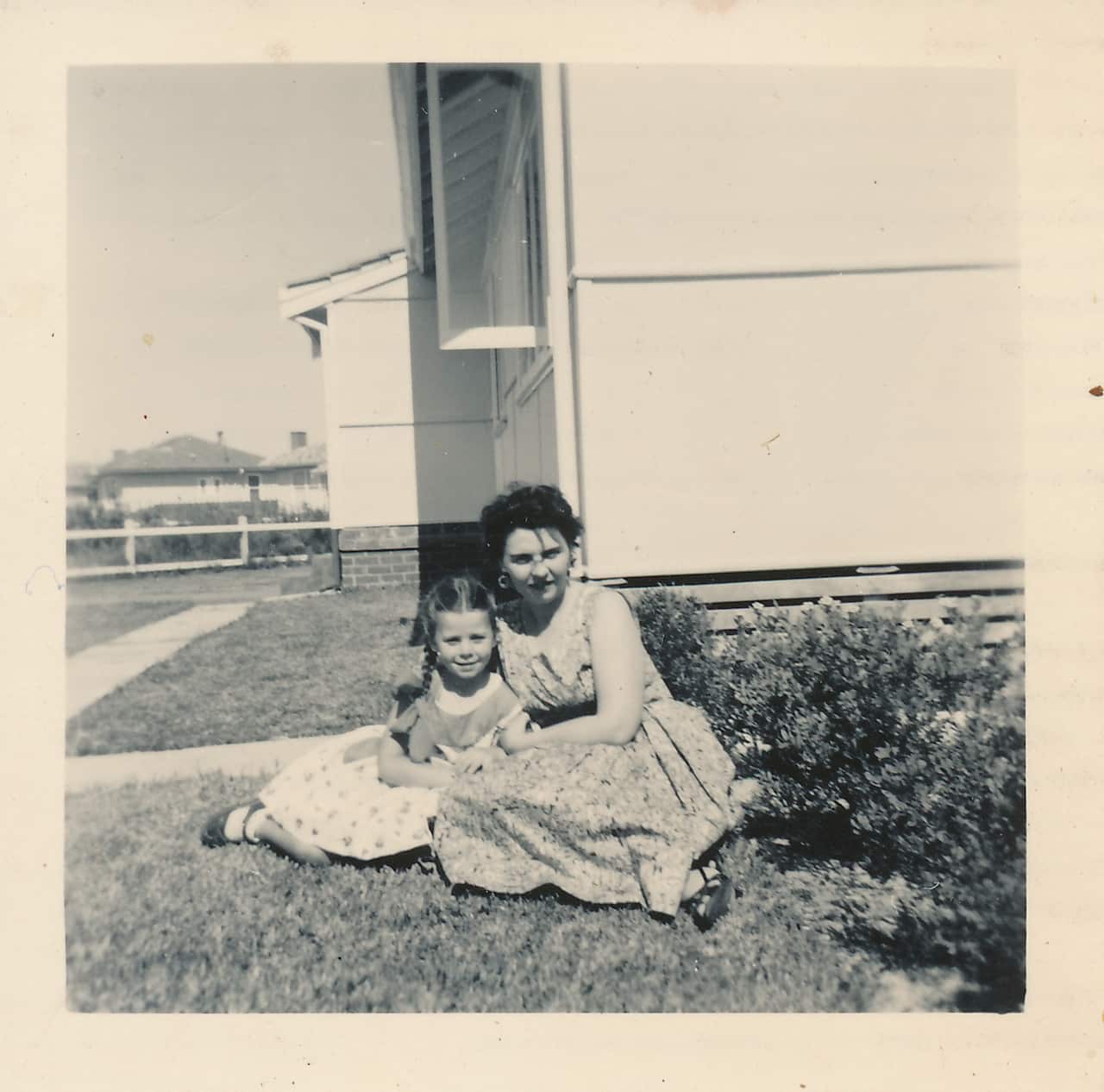 A black and white picture of a woman sitting on the grass of a suburban home cuddled up with a young girl.