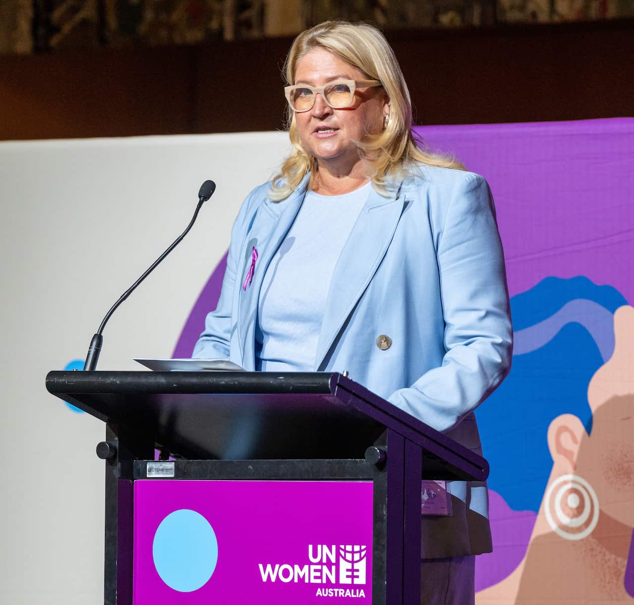 A woman in a light blue dress and a matching blazer is speaking at a UN Women Australia podium.