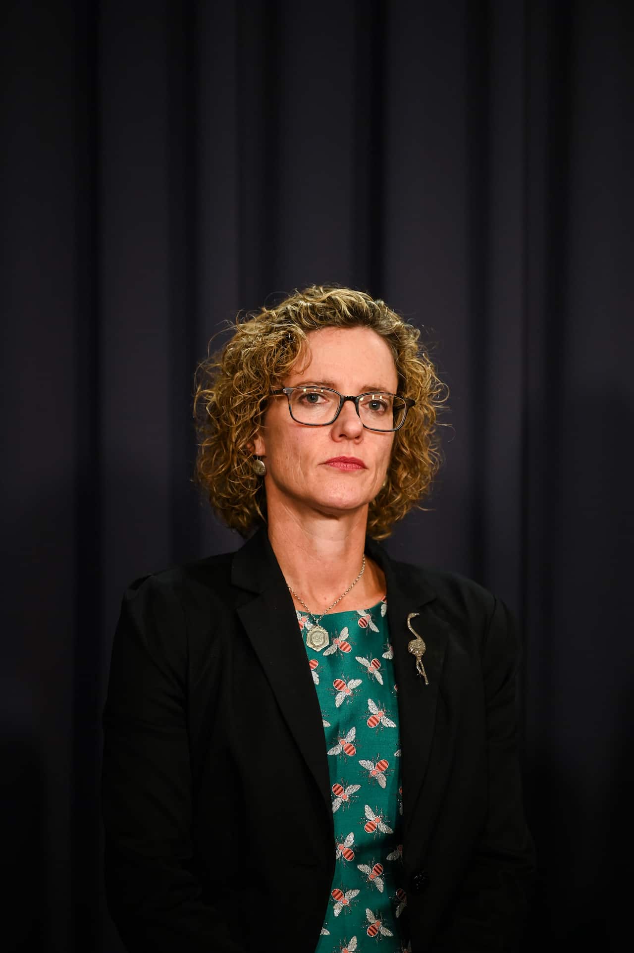 A woman wearing a green dress and dark blazer standing in front of a dark curtain.