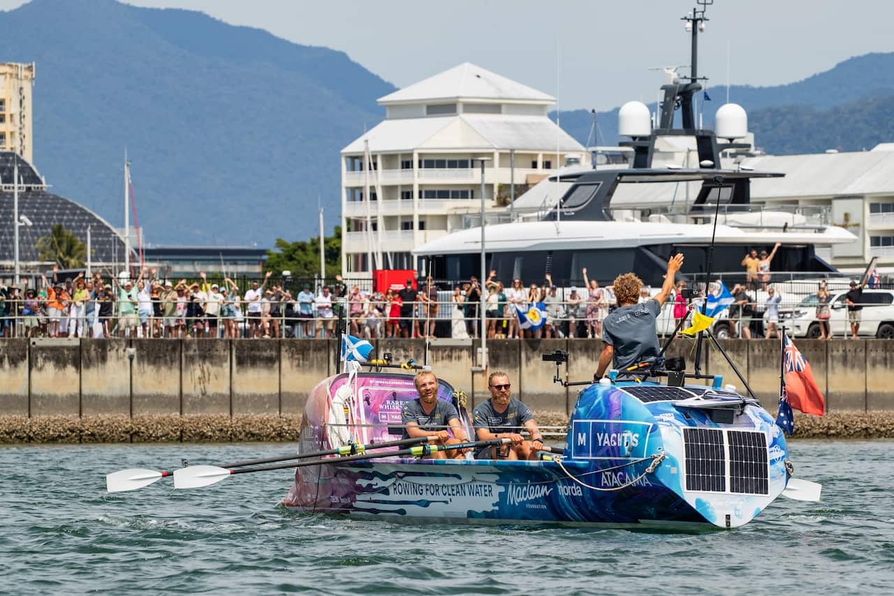 A blue rowing boat is in the water at a pier with a crowd full of people waving.