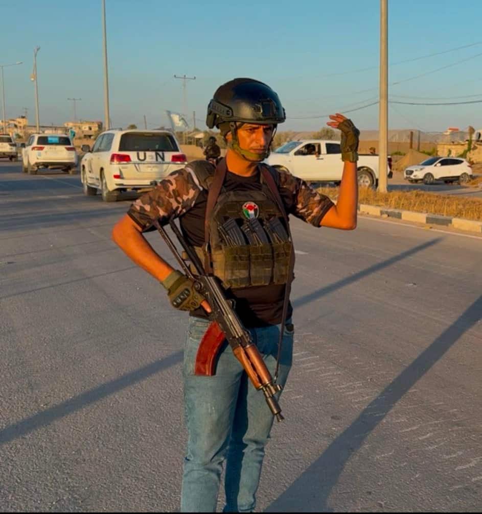 A man in a helmet and military vest holding a gun while standing on a road