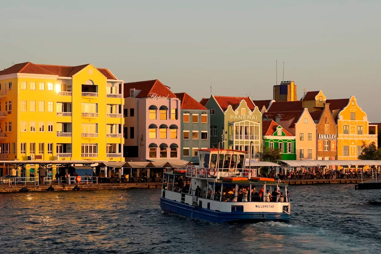 A row of colourful buildings stands along a port as a ferry approaches. Sunset casts a golden glow over the buildings. 
