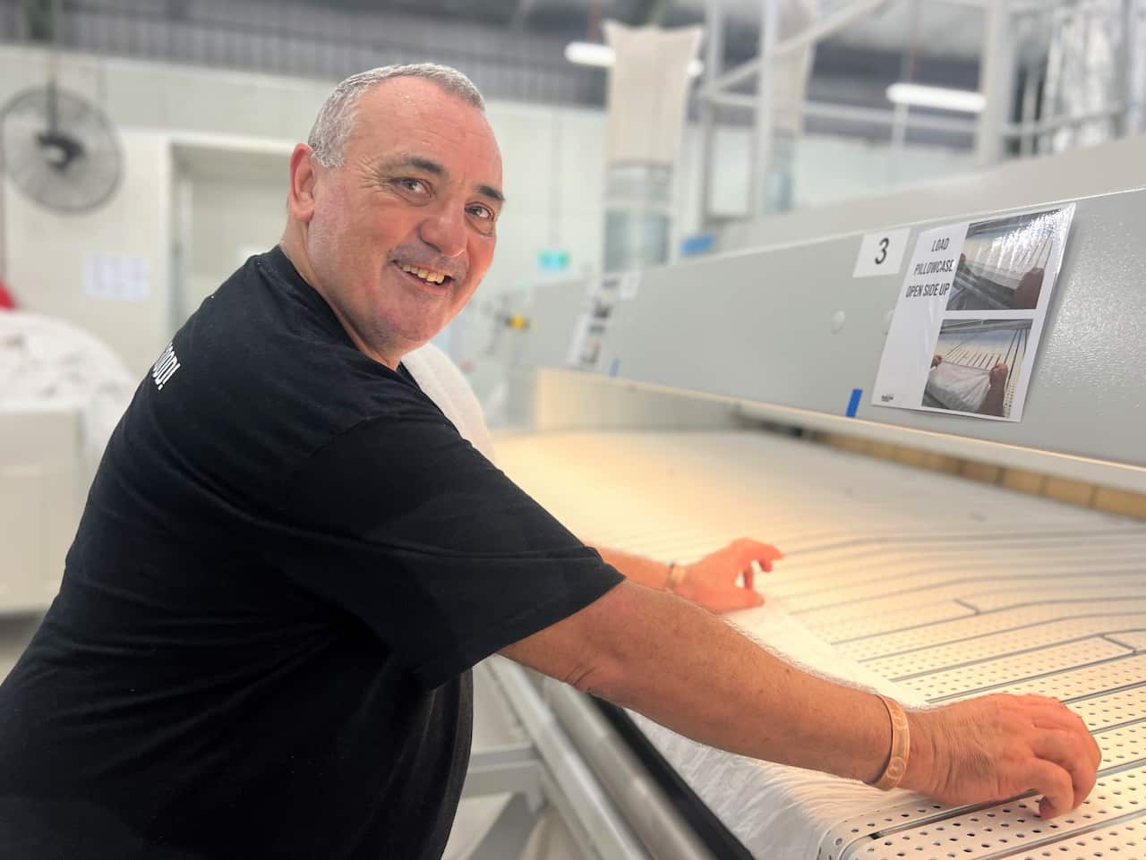 A man in a black t-shirt leans over a counter in a commercial laundry warehouse.
