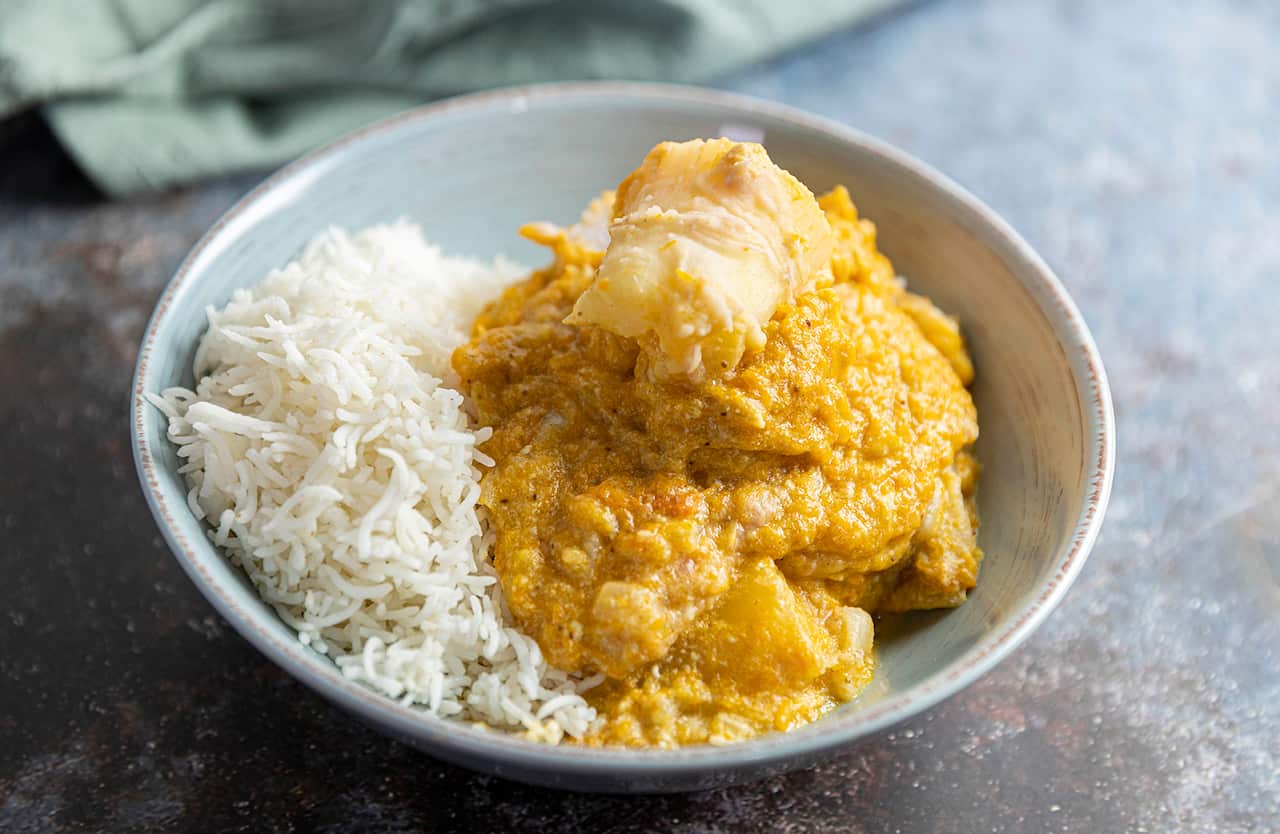A thick yellowish stew with large chunks of vegetable sits in a wide bowl, along with cooked white rice. 