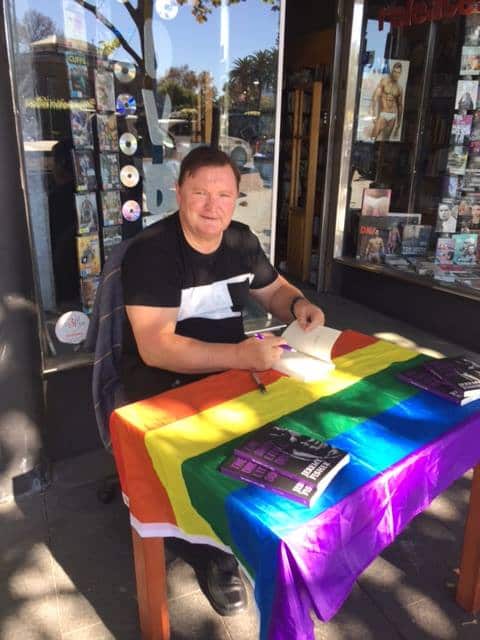 A man sitting at an outside table that is covered in a rainbow flag, with four closed books on it. He is signing a fifth book.