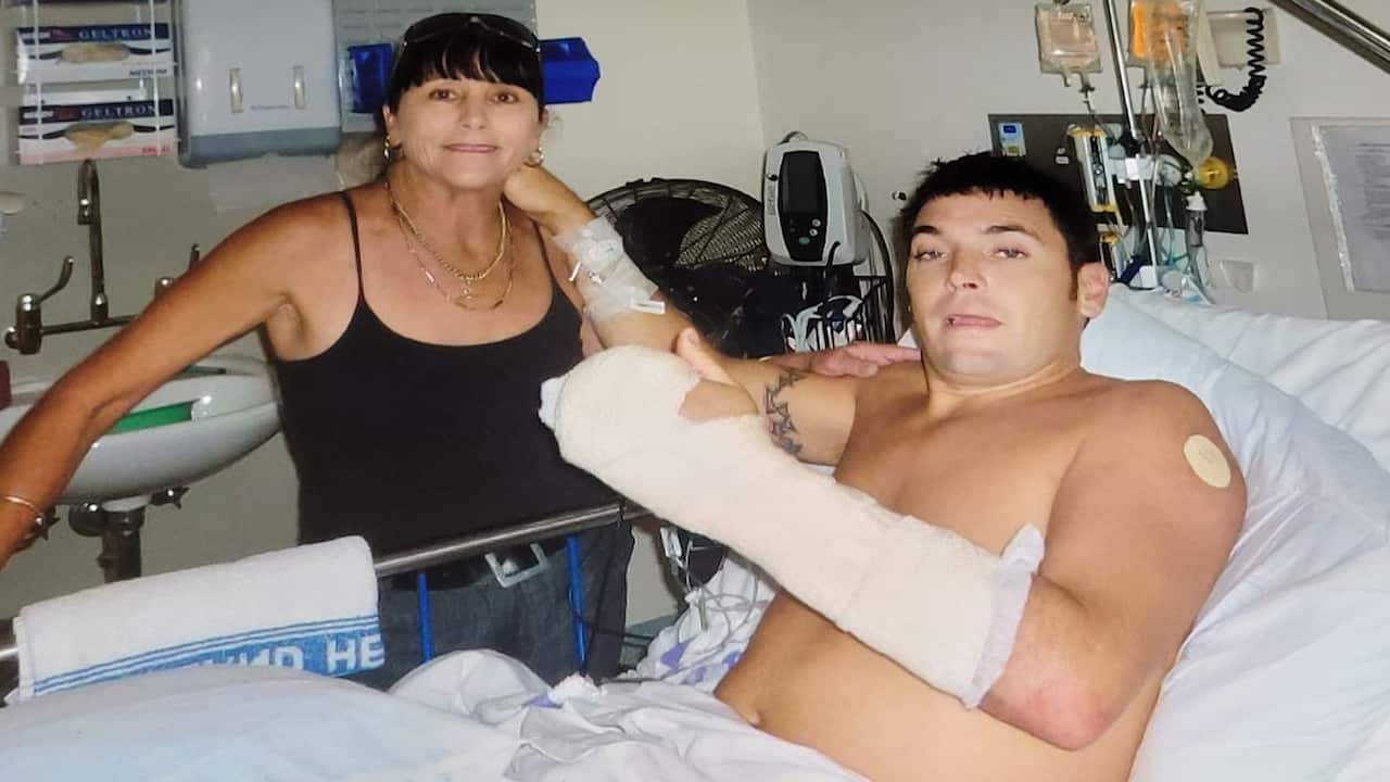 a middle aged woman with a black fringe and in a black singlet top visits a young man in hospital who has his left arm in a cast and no shirt on