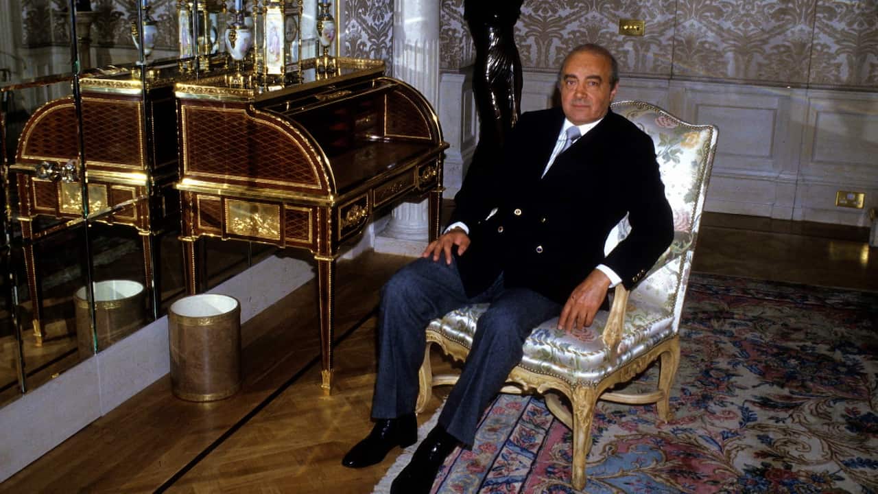 A middle-aged man wearing a black suit is sitting in an ornate vintage chair in an opulent room decorated with patterned carpet and wallpaper, gold-rimmed furniture, and mirrors