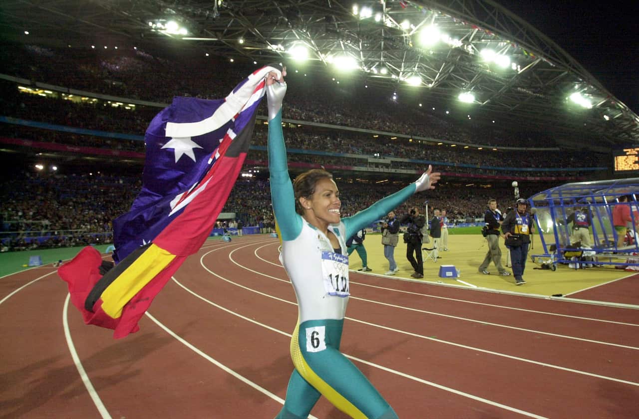 Cathy Freeman wearing a green running suit holds up the Aboriginal and Australian flags on the athletics track