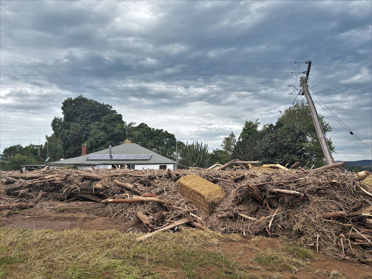 A huge pile of debris including branches, sticks and a hay bale, pushed up against a house. The pile stretches wider than the entire house and is nearly as tall. A power pole leans precariously to one side over the home.
