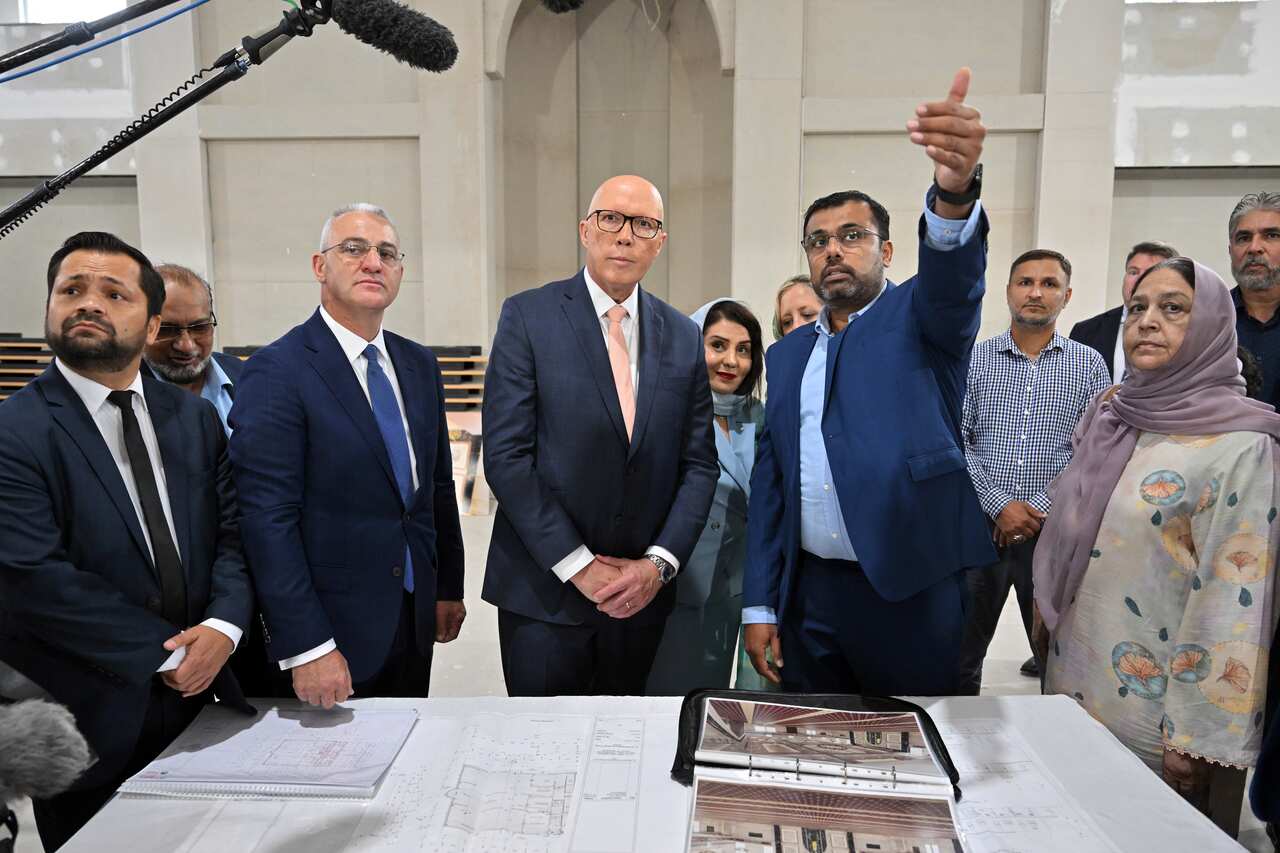 A man in a suit, hands clasped in front of him, is being directed to look at something by another man who is pointing. A group of people stands around them.