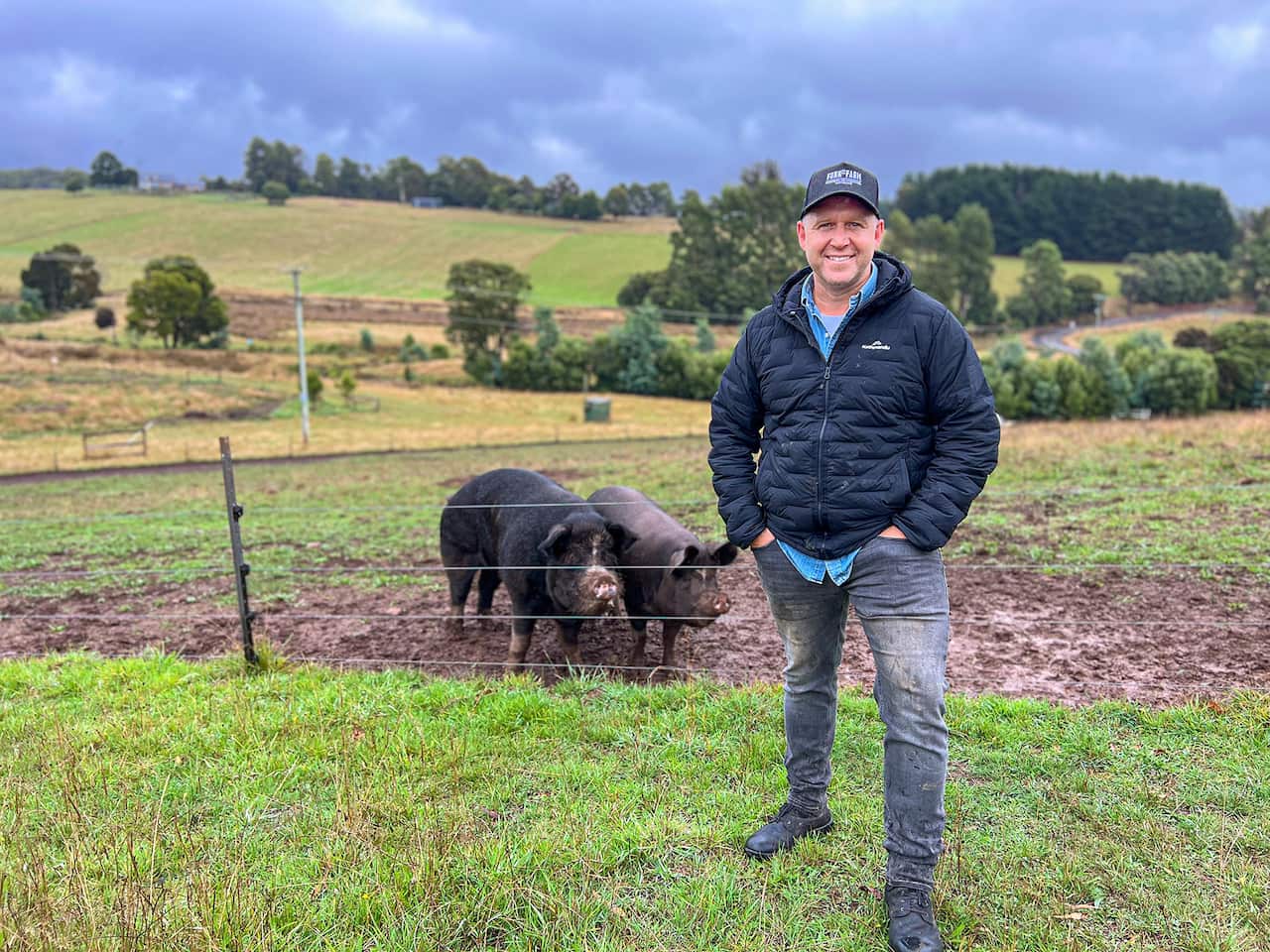 Massimo Mele stands in a farm, in front of a fence. Two black pigs are standing behind the fence. Rolling hills and blue sky can be seen in the background. 