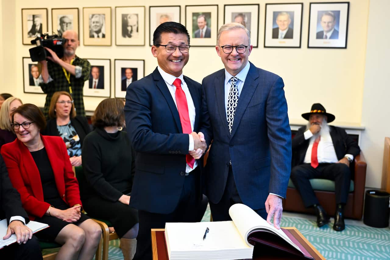 Anthony Albanese (right) shakes hands with another man who is standing next to him. Both men are wearing suits. There is a large open book with a pen resting on it on a table in front of them. There are other people sitting in the room.