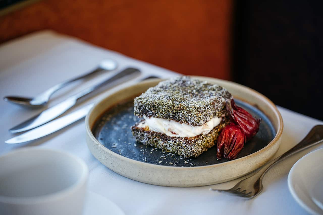 A large lamington, with yellowish flecks in the coconut coating and a generous whipped cream filling, sits on a table in a stylish round bowl, which also holds two rosella flowers.