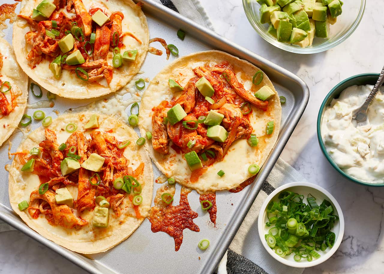 A metal baking tray sits on a marble surface. Three open tortillas, and a little of a fourth, sit on the tray, each topped with a saucy chicken mix and cubes of avocado. Bowls with sour cream, avocado and sliced green onion sit alongside the tray.
