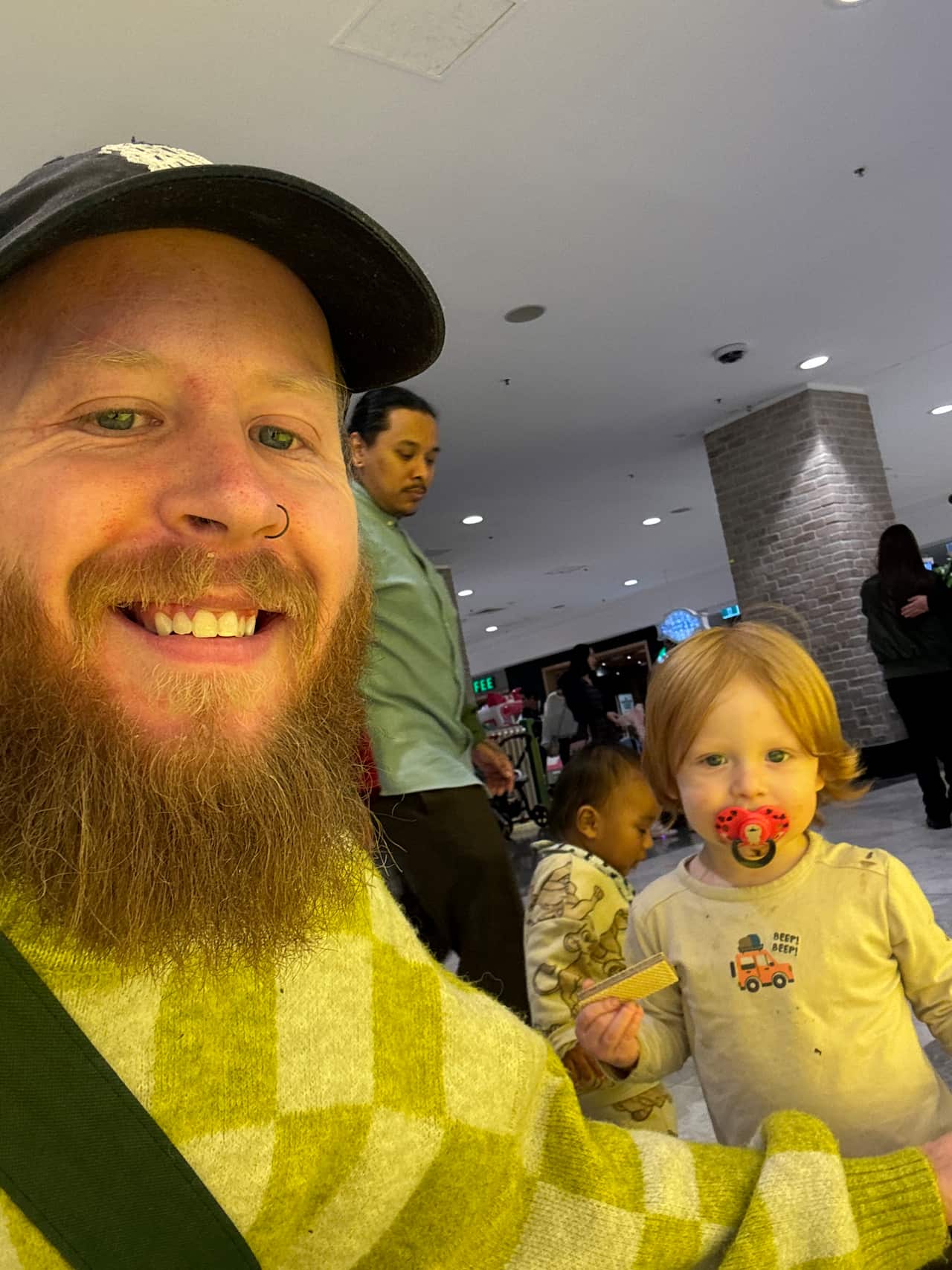 A man with a beard smiles at the camera. There is a child next to him with a dummy in its mouth.