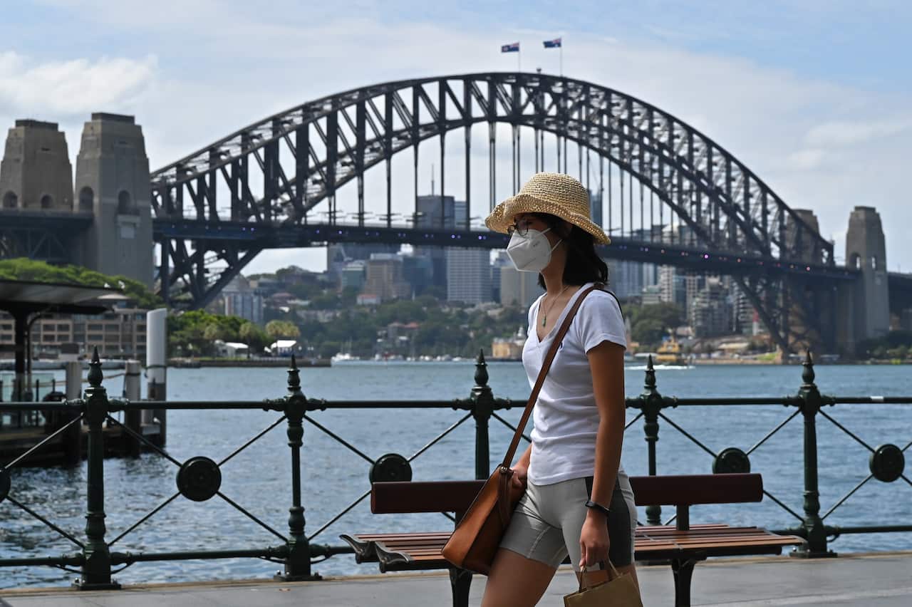 A woman wearing a hat and a face mask is walking beside Sydney harbour with the Harbour Bridge in the background.