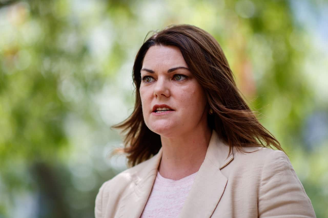 A woman in a light brown blazer standing in front of trees.
