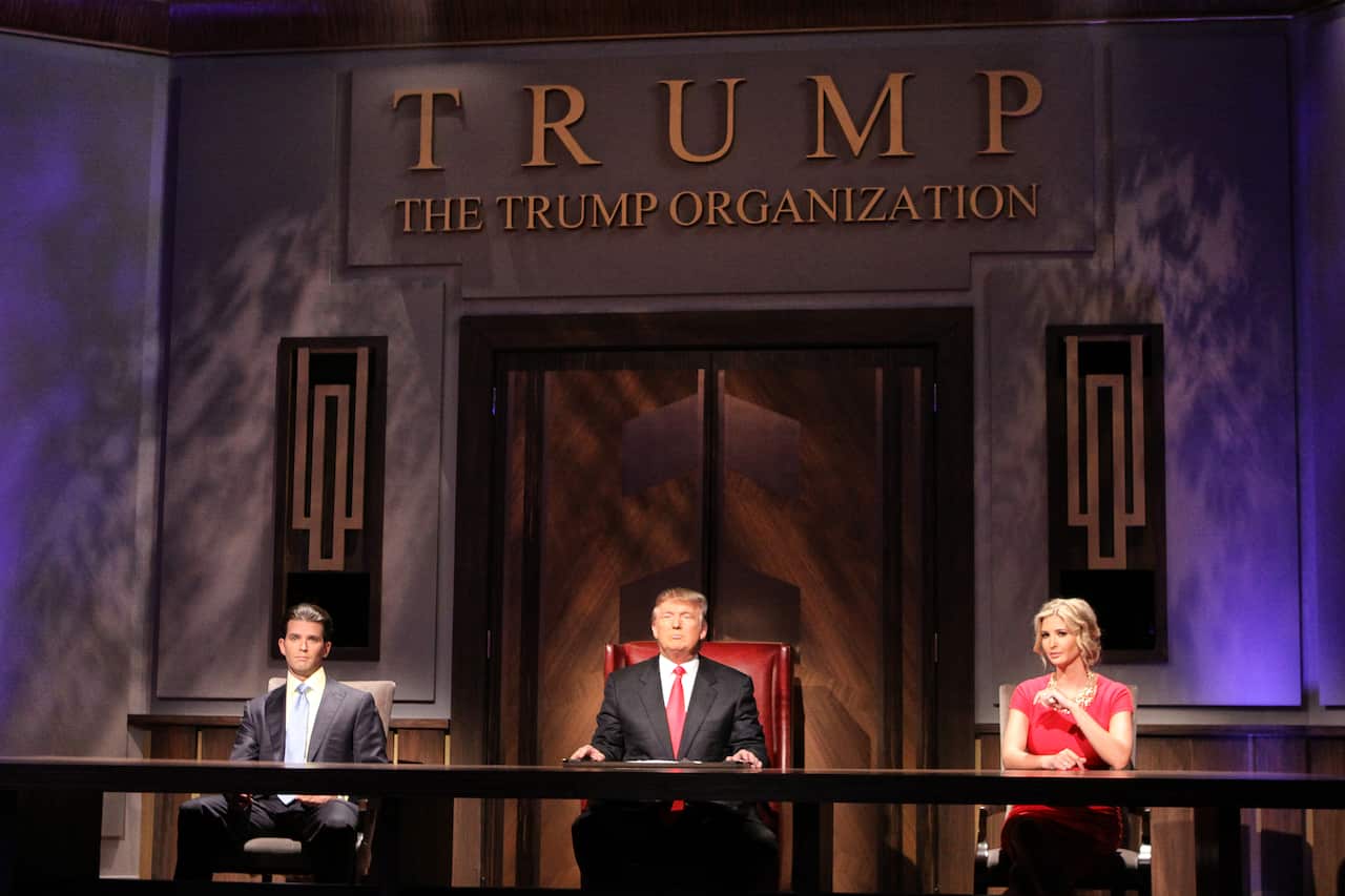 Donald Trump sits in the centre, flanked by Donald Trump Jr. on the left and Ivanka Trump on the right, in front of a gate inscribed with the words Trump The Trump Organization above it.