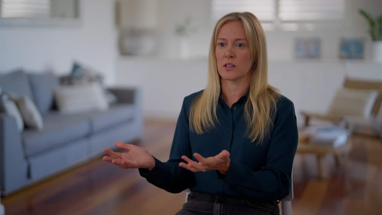 A blonde woman wearing a dark blue shirt is gesticulating while speaking