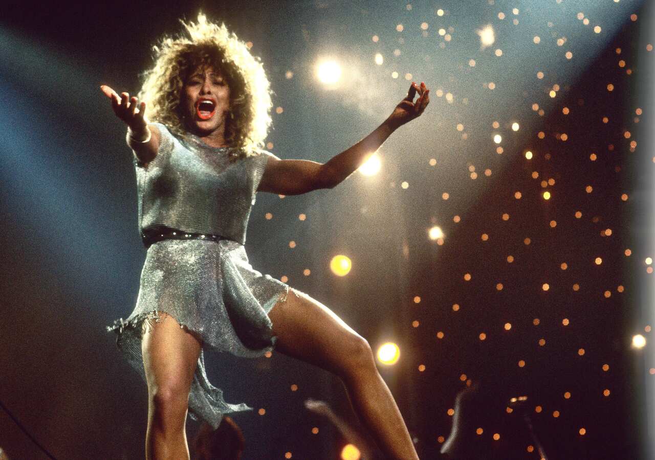 Musician Tina Turner performs on stage in a sparkly silver dress with her hands outstretched.