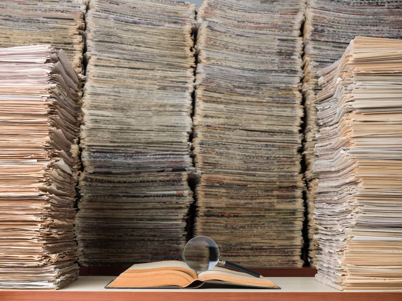 An open book with a magnifying glass on it surrounded by stacks of ducuments and newspapers.