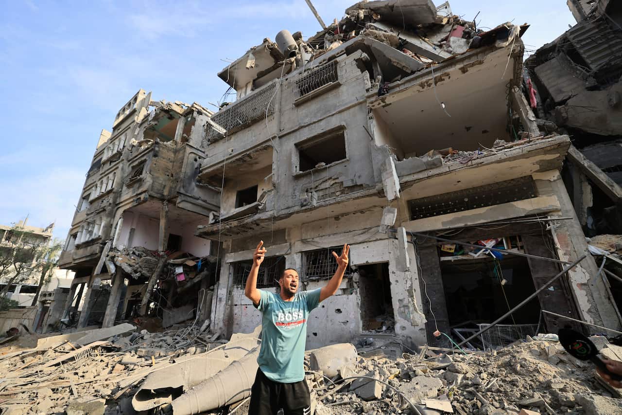 Man stands on top of rubble, throwing his arms into the air and yelling in front of destroyed buildings.
