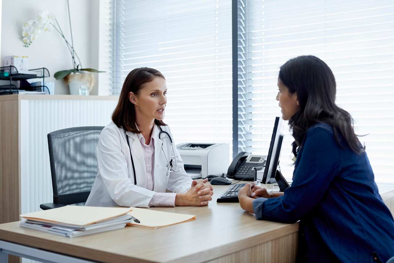 A female doctor talks with a female patient in the clinic. Both women are seated across a table, facing each other as they converse.