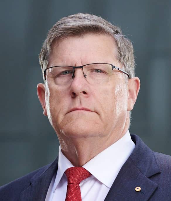 A man with short grey hair, wearing glasses, a blue blazer, white shirt and red tie. He has a neutral expression.