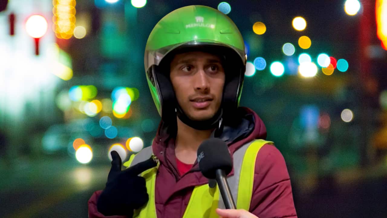 A delivery rider wearing a lime green helmet, a maroon hoodie and a yellow hi-vis vest against a blurred city background.