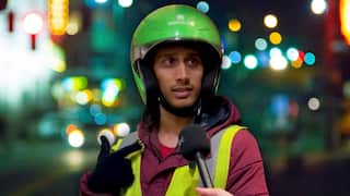 A delivery rider wearing a lime green helmet, a maroon hoodie and a yellow hi-vis vest against a blurred city background.